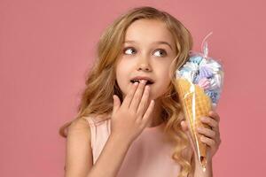 pequeño niña con un rubio Rizado cabello, en un rosado vestir es posando con un helado foto