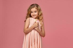 Little girl with a blond curly hair, in a pink dress is posing with a candy photo