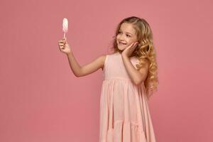 pequeño niña con un rubio Rizado cabello, en un rosado vestir es posando con un caramelo foto