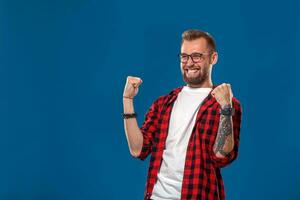 Trendy man in checkered shirt holding fists up in gesture of success and screaming happily with win on blue background photo