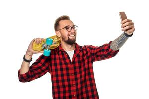 Young man holding the skateboard on shoulder raising his phone takes a selfie on a white background. photo