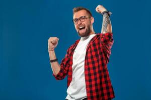 Trendy man in checkered shirt holding fists up in gesture of success and screaming happily with win on blue background photo