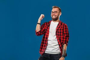 Trendy man in checkered shirt holding fists up in gesture of success and screaming happily with win on blue background photo