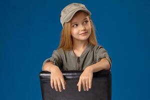 pequeño niña en un gorra con gris ojos y rubio pelo sentado en un silla foto