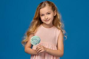 Fashionable little girl in a pink dress, with a candy in her hands, standing on a blue background. photo