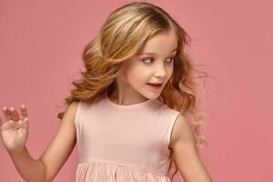 pequeño niña con un rubio Rizado cabello, en un rosado vestir es posando para el cámara foto