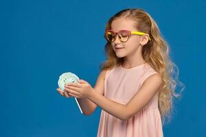 Fashionable little girl in a pink dress and glasses, with a candy in her hands, standing on a blue background. photo