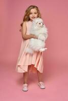 Little girl with a blond curly hair, in a pink dress plays with her dog photo