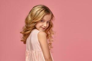 pequeño niña con un rubio Rizado cabello, en un rosado vestir es posando para el cámara foto