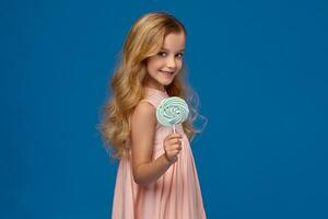 Fashionable little girl in a pink dress, with a candy in her hands, standing on a blue background. photo