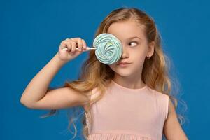 Fashionable little girl in a pink dress, with a candy in her hands, standing on a blue background. photo