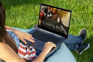 mujer trabajando en ordenador portátil ordenador personal computadora con con su culinario Blog, sentado en parque en verde césped Brillo Solar césped al aire libre. foto