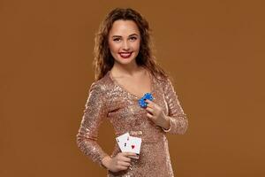 Attractive brown-haired caucasian young woman in golden cocktail dress at casino holds pair of aces and chips in her hands photo