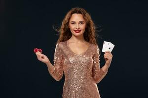 Brown-haired attractive caucasian young woman in golden cocktail dress at casino holds pair of aces and chips in her hands photo