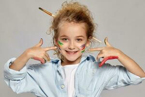 hermosa pequeño niña con un pintado manos y las mejillas es posando en un gris antecedentes. foto