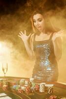 Young beautiful woman poses near poker table in luxury casino. Passion, cards, chips, alcohol, dice, gambling, casino - it is as female entertainment. Smoke background. photo