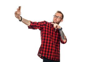 estilo de vida concepto un joven hombre con un barba en camisa participación móvil teléfono y haciendo foto de él mismo mientras en pie en contra blanco antecedentes.