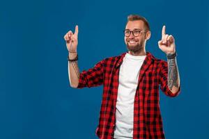 Young smiling bearded man in a checkered shirt showing direction with finger. Isolated on blue background photo