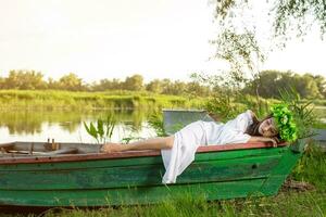 The nymph with long dark hair in a white vintage dress sitting in a boat in the middle of the river. photo