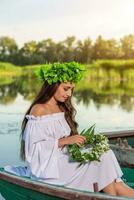 el ninfa con largo oscuro pelo en un blanco Clásico vestir sentado en un barco en el medio de el río. foto