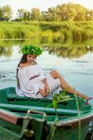 el ninfa con largo oscuro pelo en un blanco Clásico vestir sentado en un barco en el medio de el río. foto