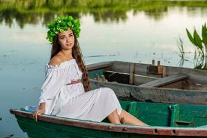 joven mujer con flor guirnalda en su cabeza, relajante en barco en río a puesta de sol. concepto de hembra belleza, descanso en el pueblo foto