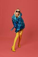 Alluring skinny young blonde woman posing in studio on red background. She is wearing blue down jacket, sunglasses, yellow stockings photo