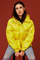Beautiful young fashion woman in yellow oversized down jacket posing in studio on red background, using chair photo