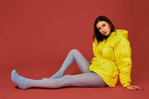 Studio portrait of young brunette woman sitting on the floor in yellow down jacket and grey blue panty hoses or stockings photo