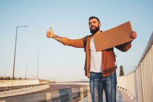 Man is hitchhiking on roadside trying to stop car. He is holding blank cardboard for your text. photo