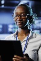 Satisfied admin in data center uses tablet to prevent system electronics overload. Efficient expert between server mainframes ensuring enough network bandwidth for smooth operations photo