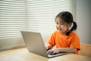 Asian baby girl wearing t-shirt smiling concentrate to use laptop and study online on wood table desk in living room at home at night. Education learning online from home concept. photo