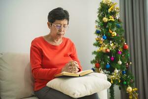 Asian old senior woman doing hands together and  holding necklace of crosses prayer to God along with the bible In the Christian concept of faith, spirituality and religion. Christmas tree. photo