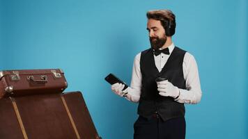 Modern doorman texting and drinking coffee in studio, enjoying work break with music on headphones. Young bellboy with professional hospitality industry job using phone and serving cold brew. photo