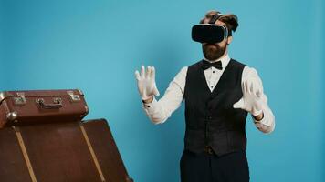 Modern doorman using vr glasses against blue background, using virtual reality technology on headset in studio. Young bellboy with classy occupation standing next to trolley bags, travel sector. photo