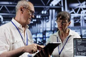 Old cloud computing business executives updating server clusters, writing intricate binary code scripts on tablet and laptop, using programming to fortify cybersecurity in high tech workplace photo