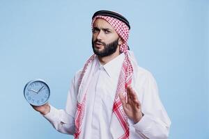 Overslept muslim man dressed in cultural clothes holding retro alarm clock and looking at camera with confused expression. Arab wearing thobe and ghutra showing time studio portrait photo