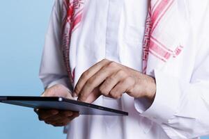 Muslim man wearing islamic traditional thobe making touch gestures with fingers on digital tablet screen closeup. Arab person using portable gadget doing zoom in on touchscreen photo