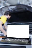 Mockup laptop on car service work station bench with repairman in blurry background replacing motor. Isolated screen device next to licensed garage specialist fixing client automobile photo