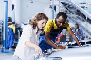 Mechanic at auto repair shop conducts annual vehicle checkup, informing customer about needed alternator replacement. Experienced garage worker talks with customer after finishing car inspection photo