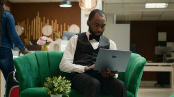 Focused hotel manager working on laptop sitting in lobby, looking at screen and keeping track of reservations to manage bookings. Bellboy in uniform works in hospitality industry. Handheld shot. photo