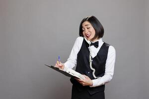 Smiling asian receptionist chatting with client on landline phone and writing in clipboard. Young attractive waitress in uniform having friendly conversation on retro telephone photo