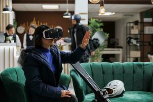 Asian woman with skiing gear engages with virtual reality device in lounge area preparing for wintersports activity. With digital VR glasses, ski resort guest balances relaxation and exploration. photo