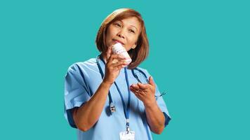 Cheerful skilled nurse holding cardiovascular bottle pill, offering medical guidance, close up. Hospital worker prescribing heart antibiotic to patient, isolated over blue studio background photo