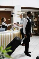Female concierge assists elderly man at front desk, providing smooth checkin process and verifying reservations. African american bellboy helps retired senior male guest with suitcase to hotel room. photo