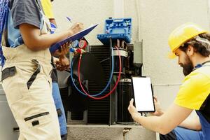 Skilled expert cleaning and lubricating outside hvac system internal evaporator coil, checking freon level and calibrating thermostats while colleagues check maintenance plan on smartphone photo