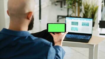 Worker looking at greenscreen display, preparing to do research for small business plan. Young adult holding smartphone with isolated copyspace mockup, open floor office space. photo