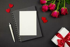 Greeting card mockup and gift box with red ribbon, hearts, roses on dark background. Valentines, mothers, womens day, anniversary and birthday greeting flat lay. Top view. Copy space. photo