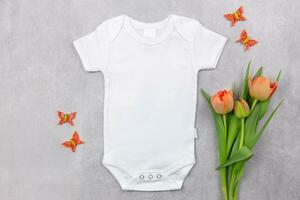 White baby girl or boy bodysuit mockup flat lay with orange tulips flowers and butterflies decoration on the gray concrete background. Design onesie template, print presentation mock up. Top view. photo