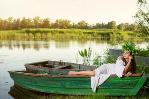 The nymph with long dark hair in a white vintage dress sitting in a boat in the middle of the river. photo